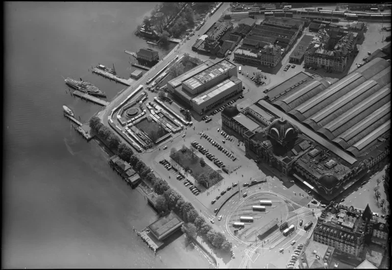 Luftaufnahme: Zwischen den Schiffsanlegern eines Sees und einem Bahnhof befindet sich eine hohe aus drei langestreckten Teilen bestehende Halle.