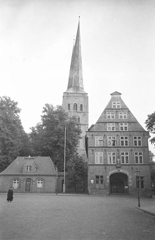 Links ein kleines Gebäude, rechts das Lübecker Zeughaus, im Hintergrund der Lübecker Dom. Im Vordergrund links läuft eine schwarz gekleidete Frau.