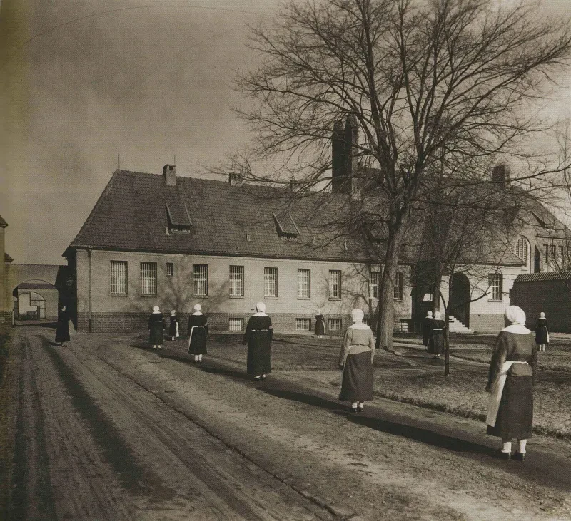 Ein Gefängnisgebäude, davor ein kahler Baum sowie zehn Frauen mit weißen Hauben und eine Frau in schwarz.