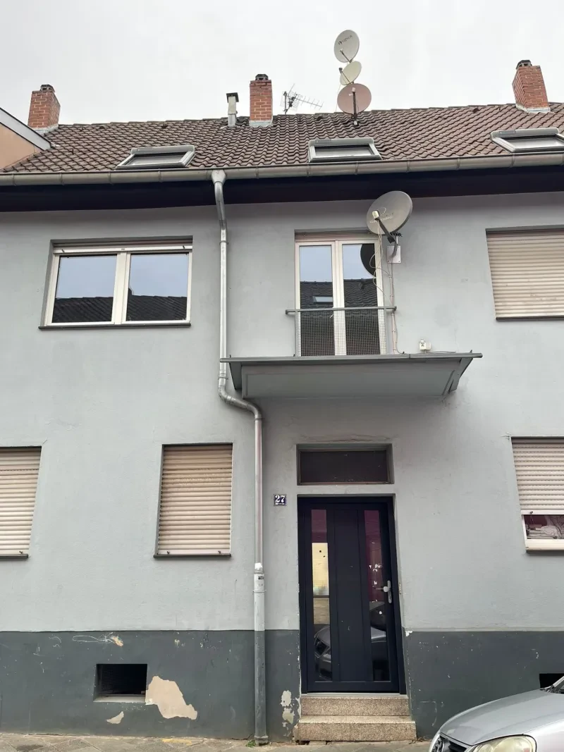Von einem zweistöckigen, grau angestrichenem Haus mit Satteldack bröckelt der Putz von der Fassade.