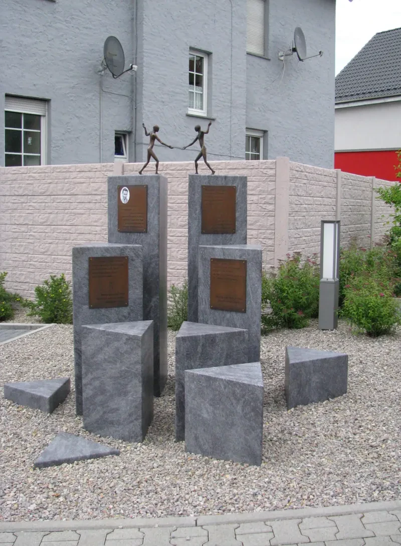 Mehrere Stelen mit Tafeln auf einem Kiesuntergrund, auf der höchsten Stele zwei Bronzefiguren.