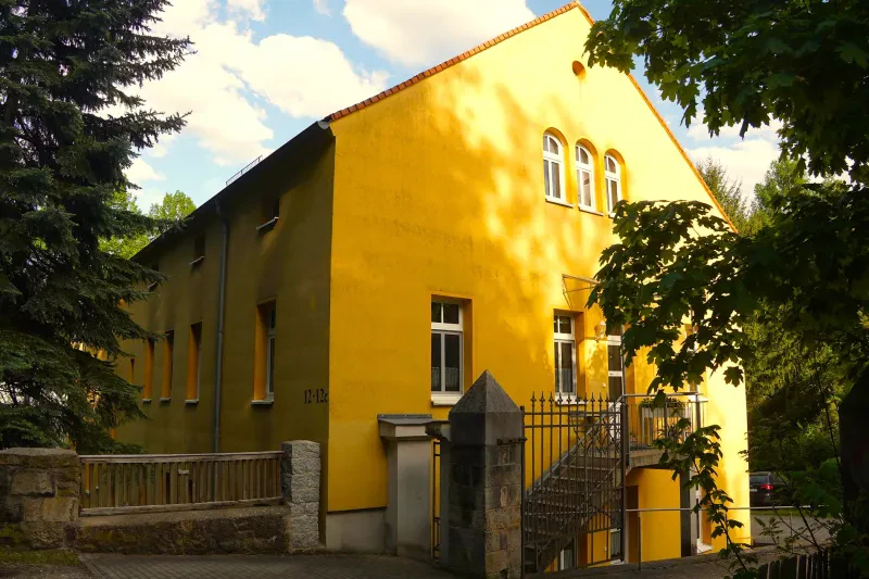 Ein gelbes, zweistöckiges Haus mit Satteldach und Außentreppe steht an einem Waldrand.