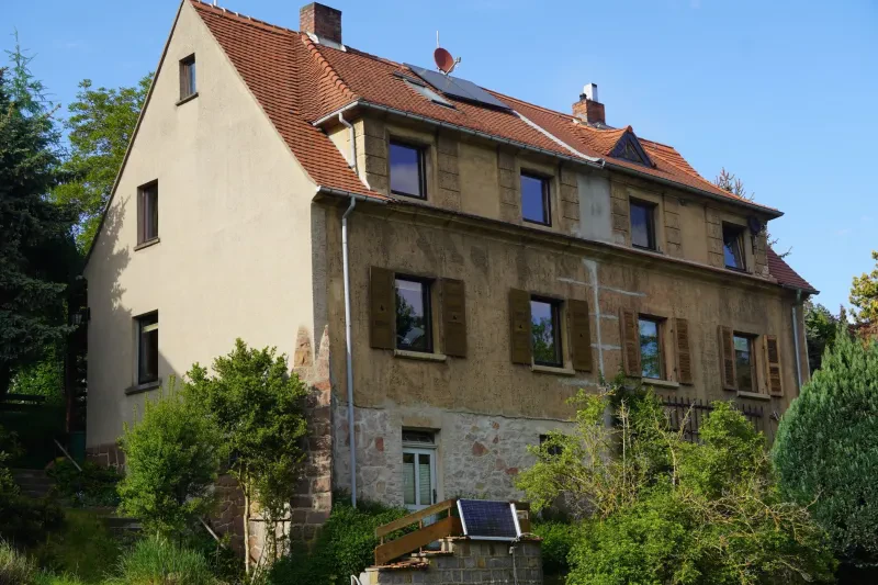 Ein dreistöckiges Wohnhaus mit Satteldach steht etwas erhöht oberhalb einer Straße am Hang. Die unterste Etage besteht aus Natursteinen, während die Etagen darüber verputzt sind.