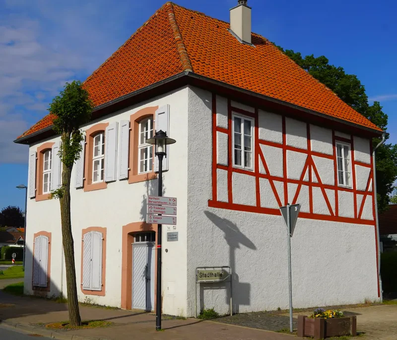 Ein kleines, zweistöckiges, weiß verputztes Gebäude mit quadratischem Grundriß und rotem Fachwerk. Die Türen und Fenster sind von ockerfarbenem Putz umgeben.
