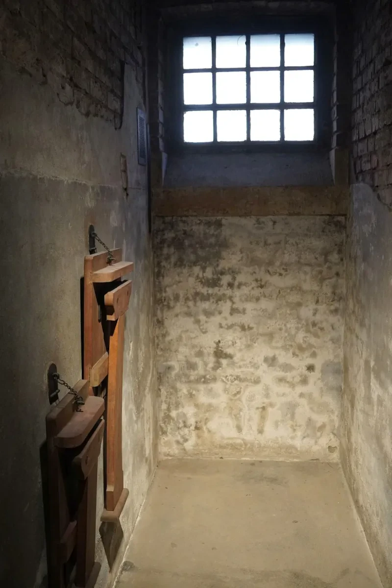 Eine schmale, dunkle Einzelzelle mit vergittertem Fenster, in der sich nur ein Klapptisch und ein Klappstuhl an der Wand befindet.