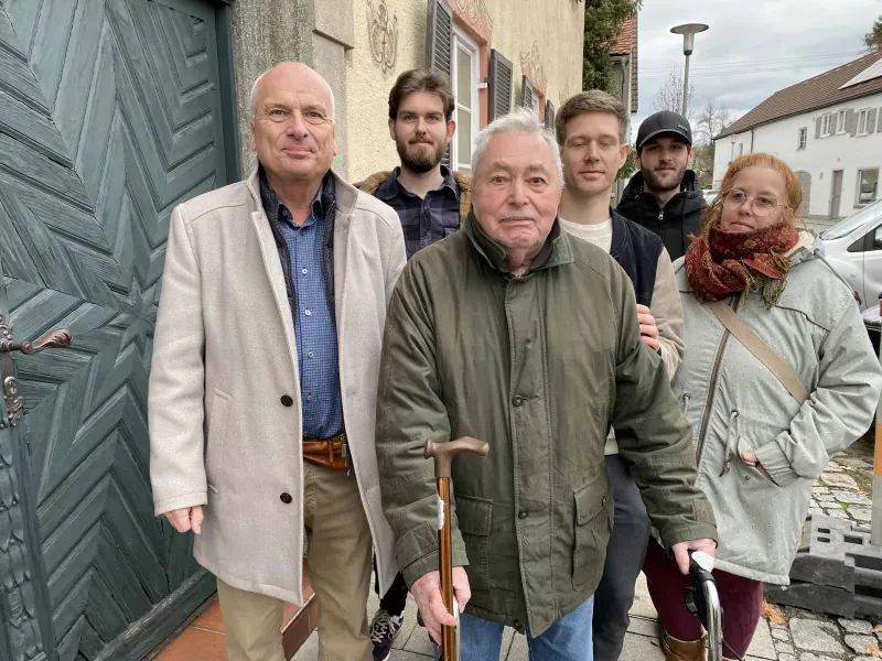 Sechs Personen unterschiedlichen Alters schauen in die Kamera. Der ältere Herr im Vordergrund stützt sich auf einen Rollator und hält einen Stock in der Hand.