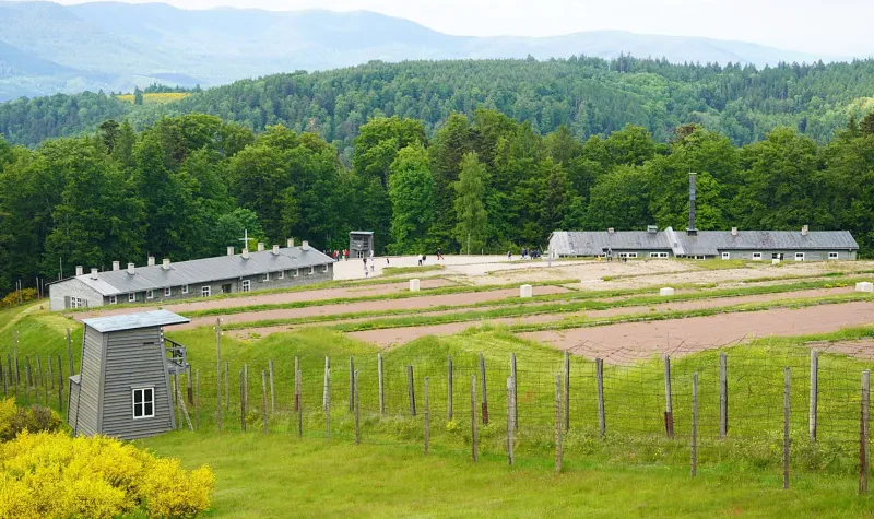 Hinter einem Zaun mit Wachturm reicht der Blick ins Tal zu zwei Baracken, dem Zellenblock und dem Krematorium.