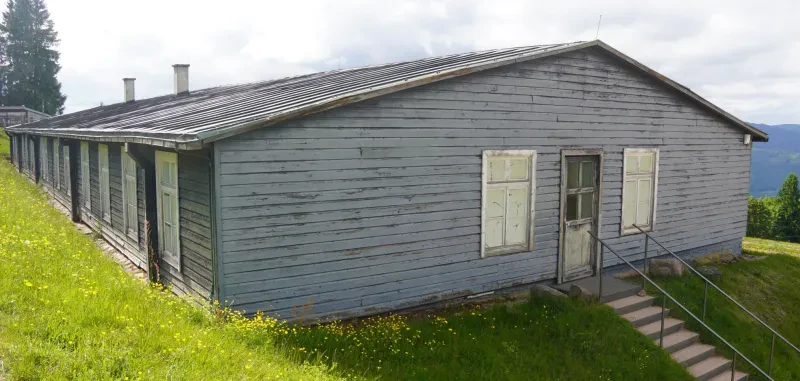 An der Längsseite einer grauen Holzbaracke befinden sich zwölf Fenster. An ihrer Stirnseite führt eine Treppe den Hang hinauf zur Eingangstür.