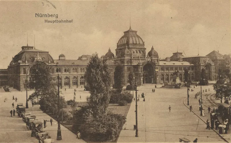 Auf einer historischen Postkarte ist ein Bahnhofsgebäude hinter einem Vorplatz zu sehen.