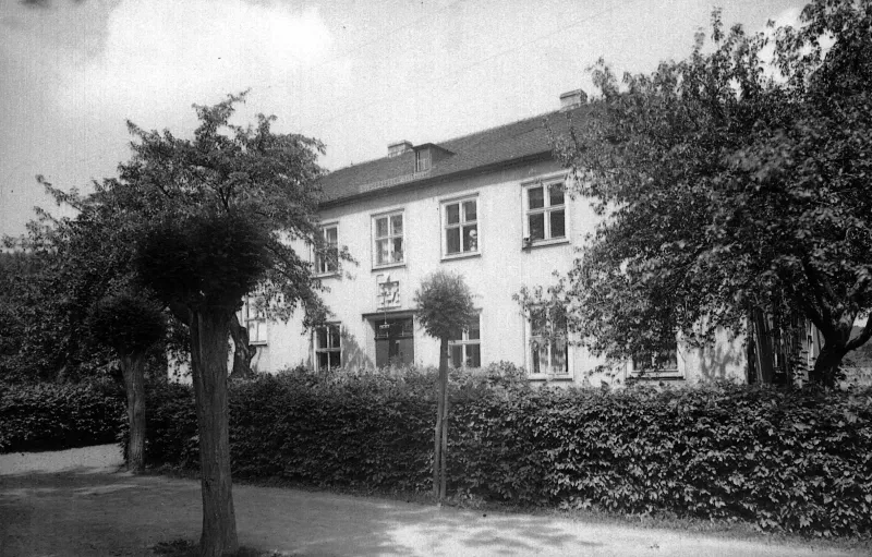 Hinter einer Hecke und Bäumen ist ein zweistöckiges weißes Gebäude zu sehen.