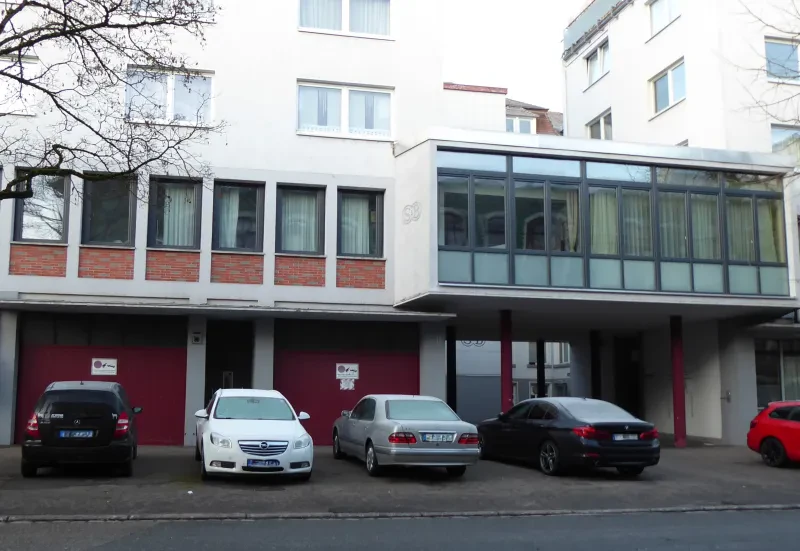 Ein Neubau, im Erdgeschoss sind Garagen, davor stehen parkende Autos.