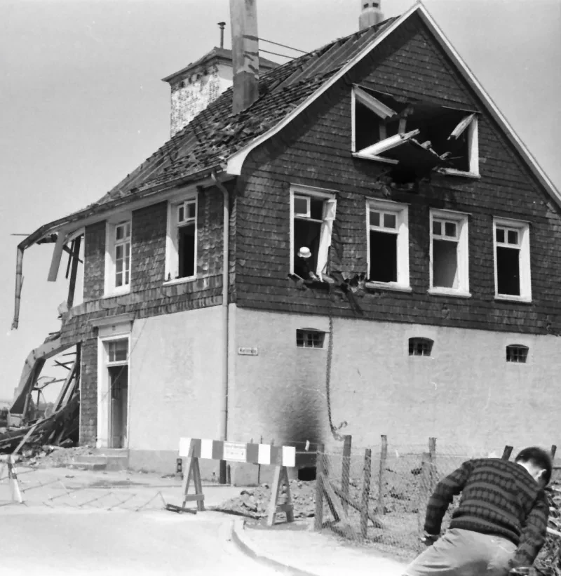 Ein Bagger ist dabei den hinteren Teil eines dreistöckigen Gebäudes abzureißen, während im vorderen Teil ein Arbeiter das Holz der Fensterrahmen aus dem Haus bricht. Die Glasfenster wurden bereits zuvor entfernt.