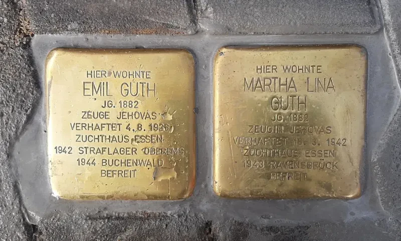 Zwei Stolpersteine liegen nebeneinander. Auf dem linken Stein steht die Inschrift: "Hier wohnte Emil Guth, Jahrgang 1882, Zeuge Jehovas, verhaftet 4. August 1936, Zuchthaus Essen, 1942 Straflager Oberems, 1944 Buchenwald, befreit." Rechts daneben liegt ein Stein mit der Aufschrift: "Hier wohnte Martha Lina Guth, Jahrgang 1882, Zeugin Jehovas, verhaftet 16. März 1942, Zuchthaus Essen, 1943 Ravensbrück, befreit". Regentropfen fallen auf die Steine.