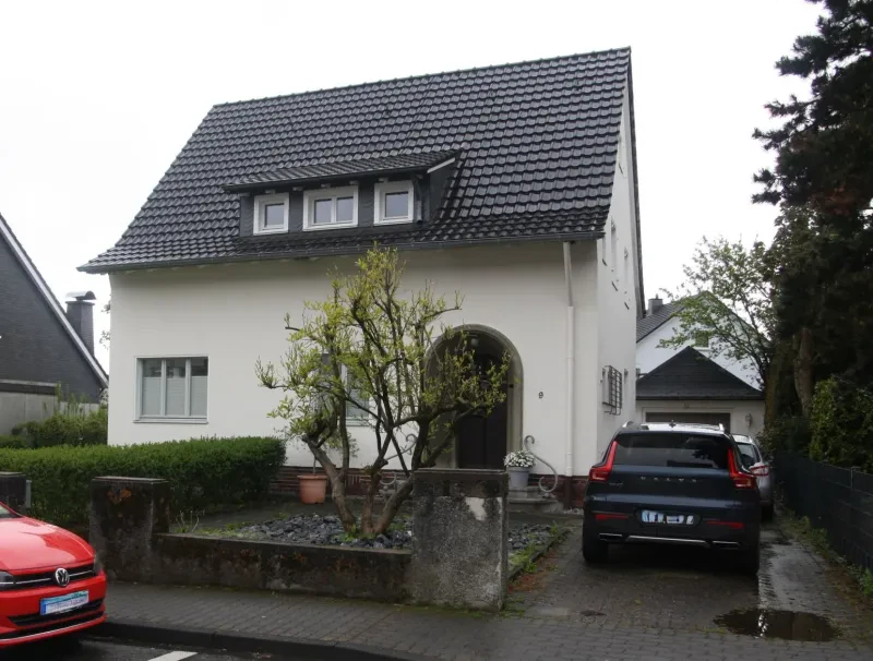 Ein weißes Einfamilienhaus mit einem Satteldach aus schwarzen Ziegeln steht auf einem Grundstück mit kleinem Vorgarten.