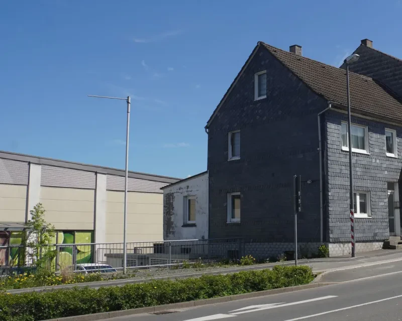 Links neben einigen schwarz verschieferten Häusern befindet sich hinter einem Geländer eine tiefergelegene Freifläche. Dahinter sieht man die Wand eines Edeka-Marktes.