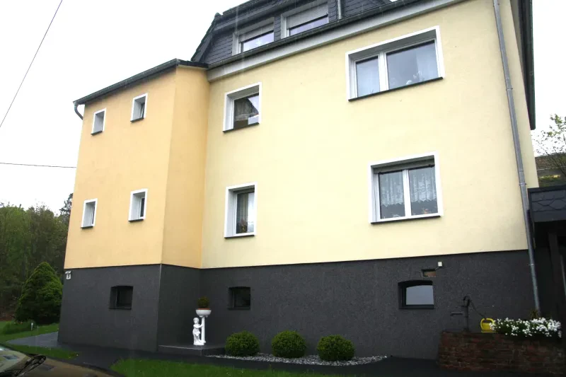 Ein dreistöckiges gelb verputztes Wohnhaus mit dunklem Sockel, dessen oberstes Stockwerk ein ausgebautes Dachgeschoß ist.