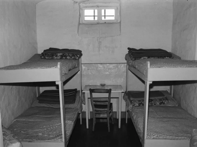 In einem engen Raum stehen links und rechts zwei Etagenbetten mit Matrazen und Decken, dazwischen ein kleiner Tisch mit einem Holzstuhl. Das kleine Fenster weit oben im Raum ist vergittert.