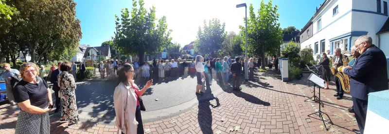 Eine große Gruppe gut gekleideter Personen steht auf der Straße vor einem zweistöckigen, weiß gestrichenen Wohnhaus.