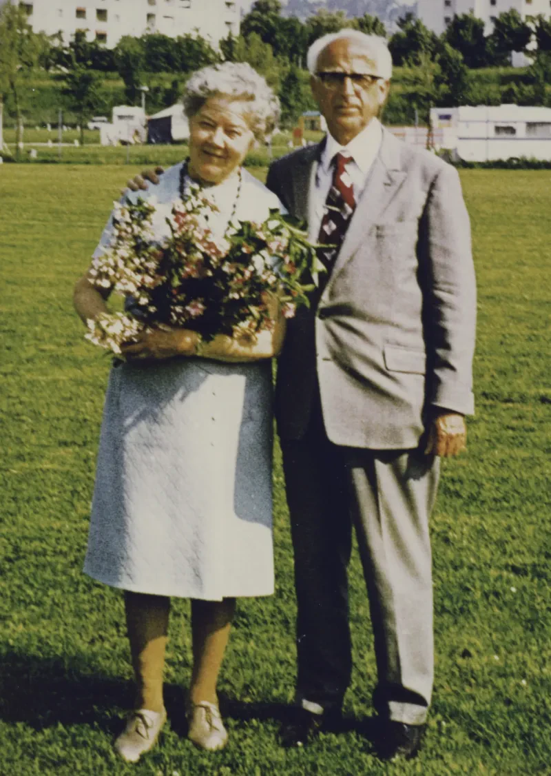 Ein älteres Paar auf einer Wiese, sie sind festlich gekleidet, sie trägt Blumen im Arm.