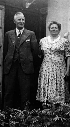 Ein älteres Ehepaar: der Mann trägt einen dunklen Anzug mit Weste, die Frau ein geblümtes Kleid.