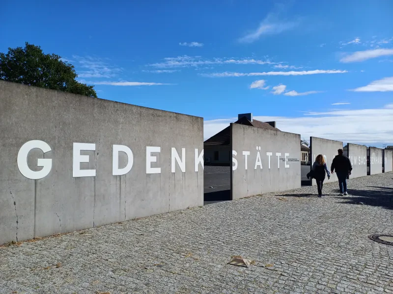 Eine Mauer mit Durchgängen, davor zwei Personen. Auf der Mauer steht: "Gedenkstätte und Museum Sachsenhausen".
