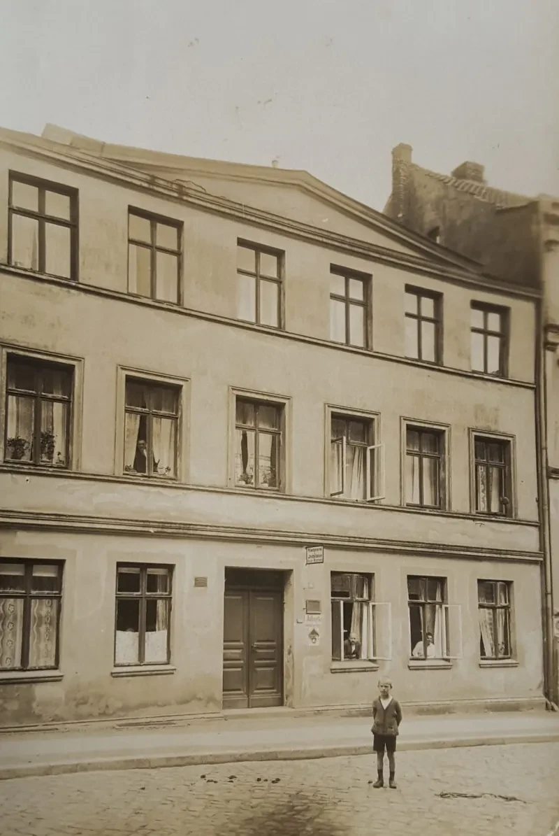 Ein dreistöckiges Wohnhaus, mehrere Personen schauen aus den Fenstern, ein kleiner Junge steht vor dem Haus.