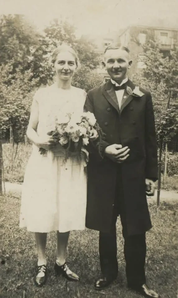 Ein junges Ehepaar am Tag ihrer Hochzeit in einem Park vor einem mehrstöckigen Haus. Die Braut hat einen Brautstrauß in der Hand und hat sich mit dem anderen Arm bei ihrem Mann eingehakt. Sie trägt ein weißes Kleid und ihr Mann einen langen, dunklen Anzug.