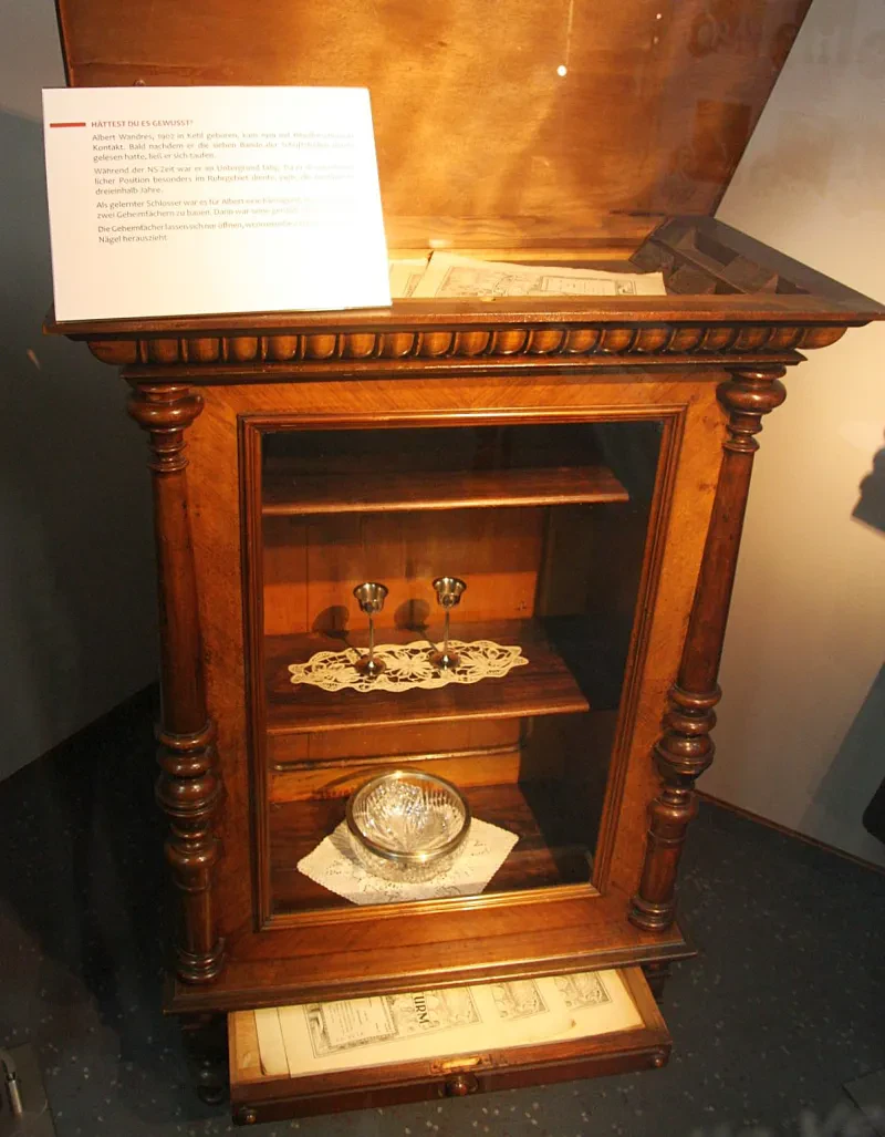 Im Boden einer Vitrine ist ein geöffnetes, ausziehbares Geheimfach mit einigen Ausgaben der Zeitschrift der Wachtturm darin. Ein weiteres, klappbares Geheimfach befindet sich im Deckel der Vitrine.
