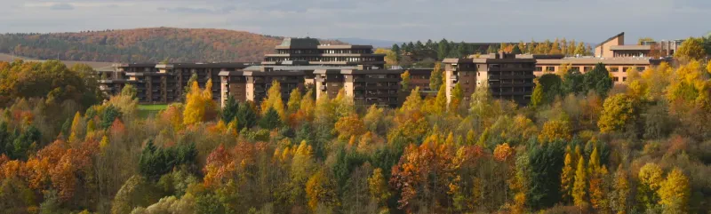 Hinter herbstlich gefärbten Laubbäumen sieht man einige Wohngebäude, die von einem Verwaltungsgebäude überragt werden. Rechts im Bild ist eine dreistöckige Druckerei zu sehen.