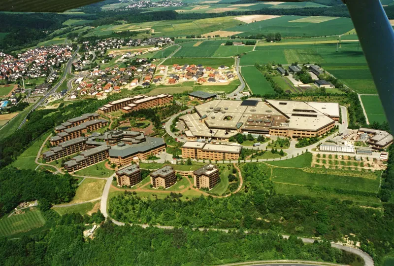 Eine Luftaufnahme zeigt einen Gebäudekomplex im Grünen, dahinter eine Ortschaft.
