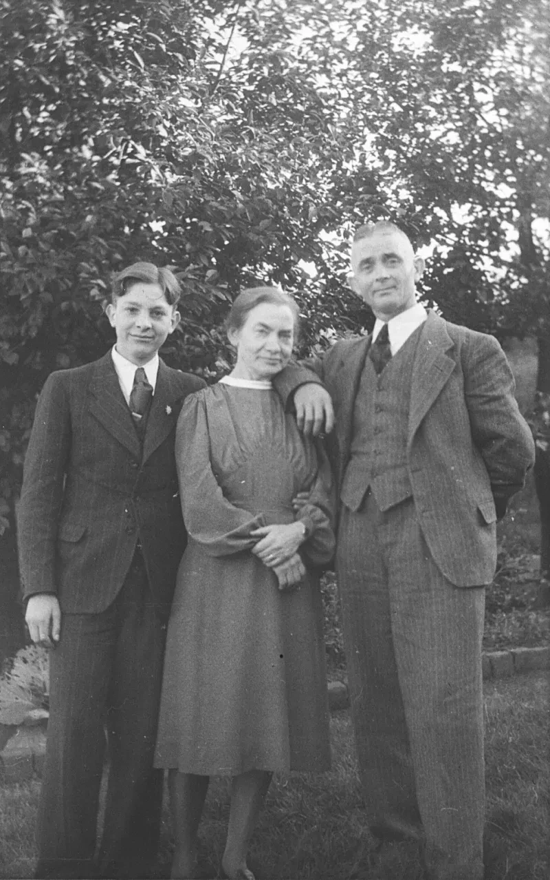 Vater und Sohn stehen im Garten und tragen einen dunklen, gestreiften Anzug. Die dazwischen stehende Mutter trägt ein Kleid.