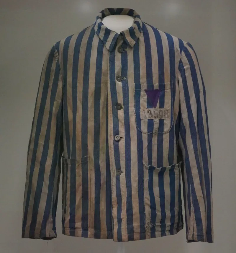 Eine blau-weiß-gestreifte Sommerjacke aus Baumwolle mit Metallknöpfen und einem aufgenähten lilafarbenen Stoffwinkel mit der Häftlingsnummer 13598.