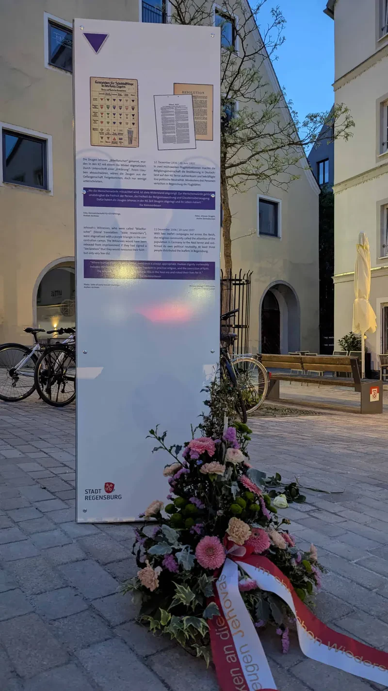 Gedenkstele mit Abbildungen und Text, davor ein Blumengebinde mit einer Schleife "zum Gedenken an die Zeugen Jehovas"