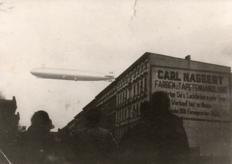 Menschen stehen vor einem Häuserblock und sehen auf einen Zeppelin, der darüber schwebt. Auf der Fassade des Häuserblocks ist eine Werbeschrift zu lesen: "Carl Naggert. Farben u. Tapetenhandlung"