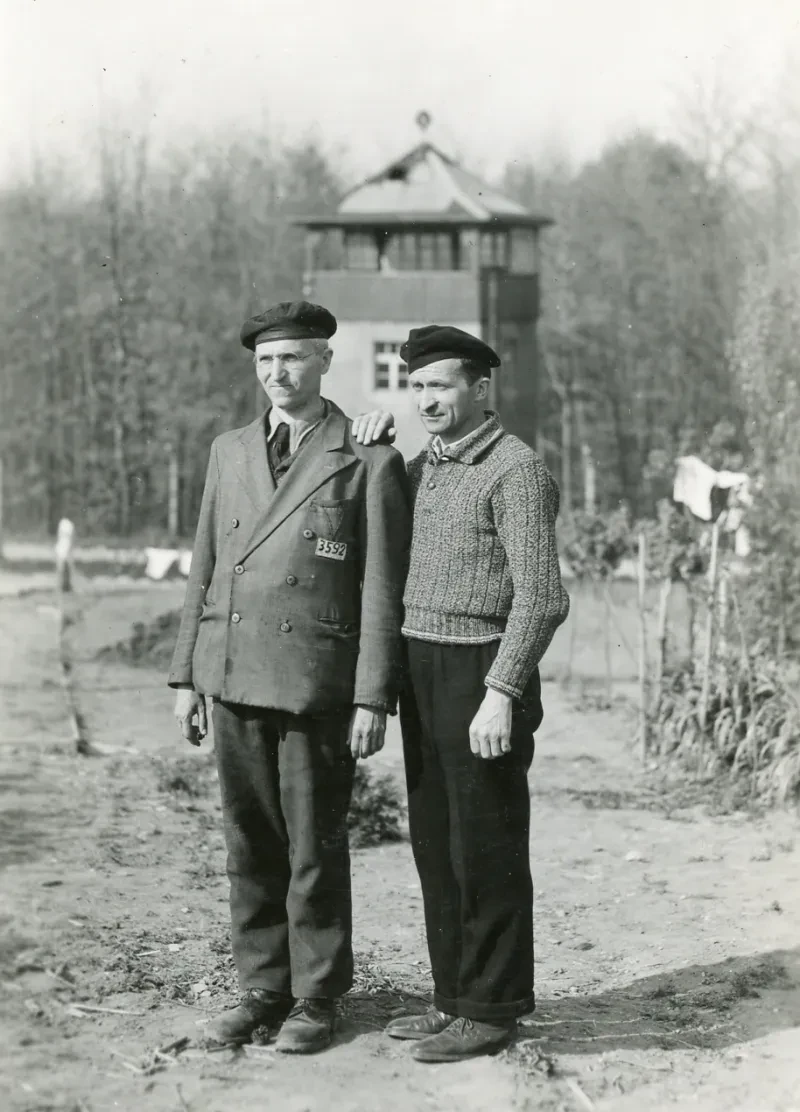 Zwei Männer vor einem KZ-Wachtturm, der linke trägt eine Jacke mit Winkel und der Häftlingsnummer 3592.