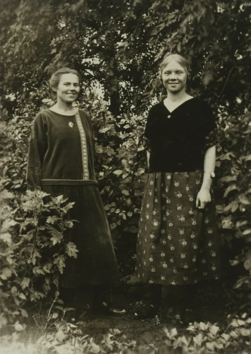 Zwei Frauen im Freien vor Bäumen und Sträuchern.