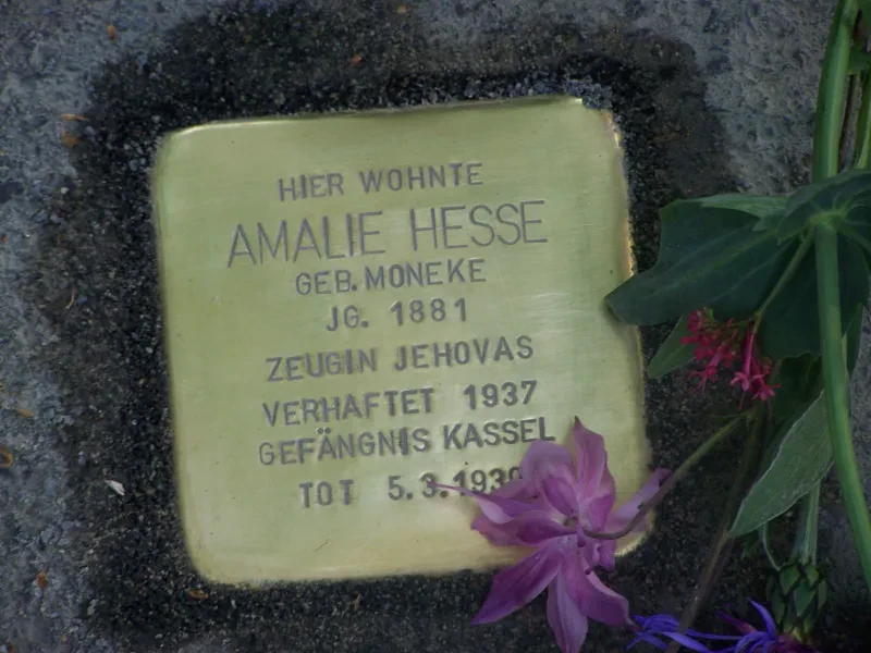 Ein Stolperstein mit der Inschrift „Hier wohnte / Amalie Hesse geb. Moneke / Jg. 1881 / Zeugin Jehovas / Verhaftet 1937 / Gefängnis Kassel / Tot 5.3.1939“ Auf dem Stolperstein liegt eine Blume.