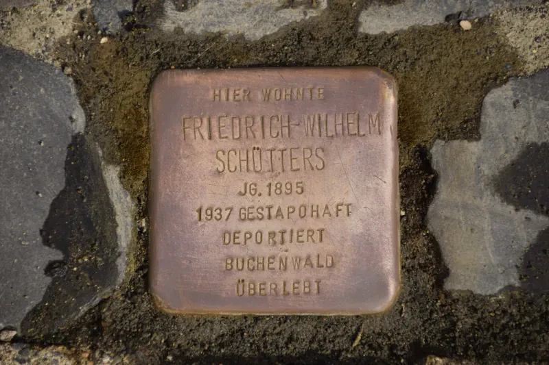 Ein Stolperstein trägt die Inschrift: "Hier wohnte Friedrich-Wilhelm Schütters, Jahrgang 1895, 1937 Gestapohaft, deportiert, Buchenwald, überlebt."