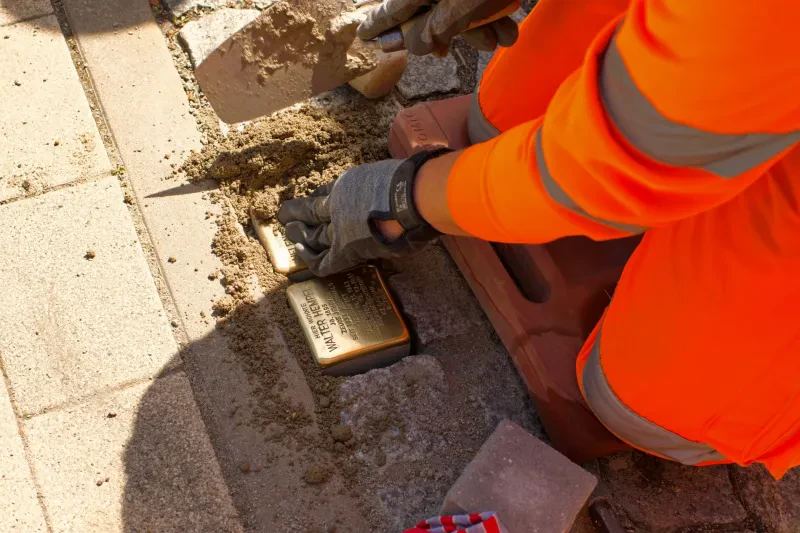 Ein in roter Signalfarbe gekleideter Mitarbeiter der Stadt fügt Sand in die Fugen zwischen den Stolpersteinen ein.