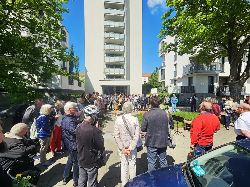 Vor einem elfstöckigen Wohnhaus stehen ca. 80 Personen in einem Kreis aufgestellt und hören einem Vortrag zur Würdigung der NS-Opfer zu.