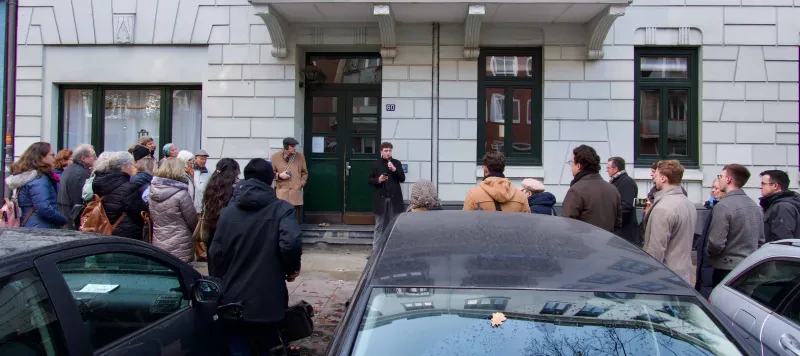Eine Menschenmenge steht vor einem Wohnhaus, davor parken Autos. Ein junger Mann steht vor dem Hauseingang und spricht in ein Mikrofon.