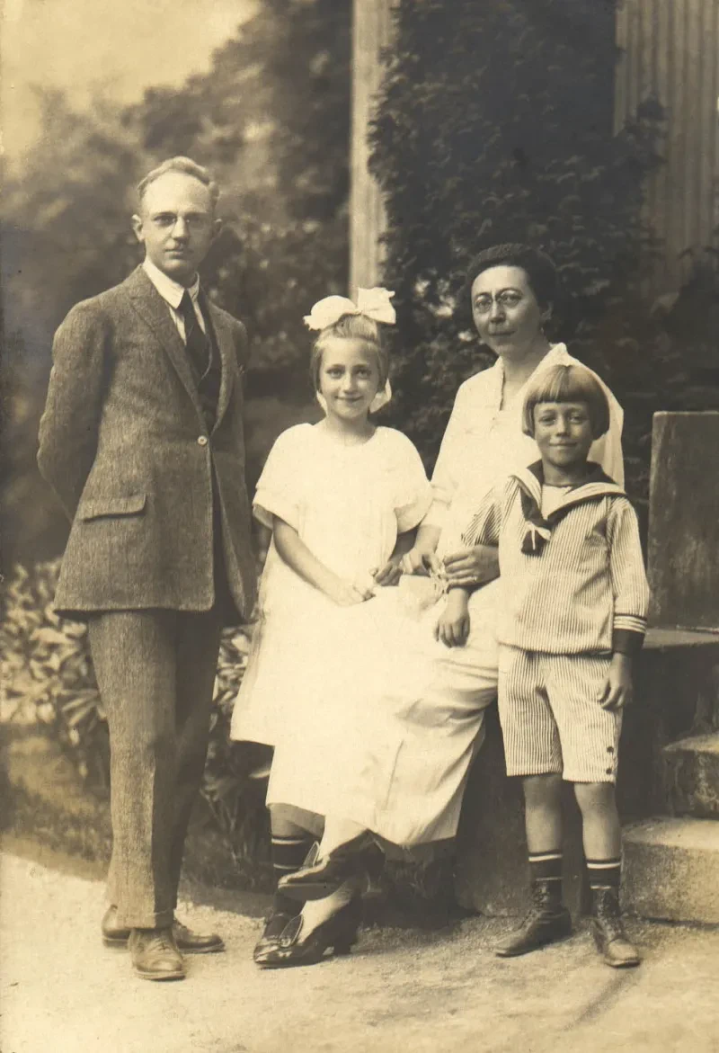 Eine Familie schaut in die Kamera. Der Vater steht links, die Mutter sitzt rechts auf einer Mauer. Vor ihr stehen ein Mädchen mit Schleife im Haar und ein Junge im Matrosenanzug.