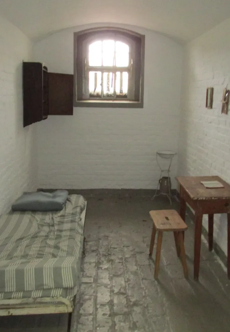 Eine Zelle mit Fenster, Wandschrank, Bett, Stuhl und Hocker und einer Waschschüssel.