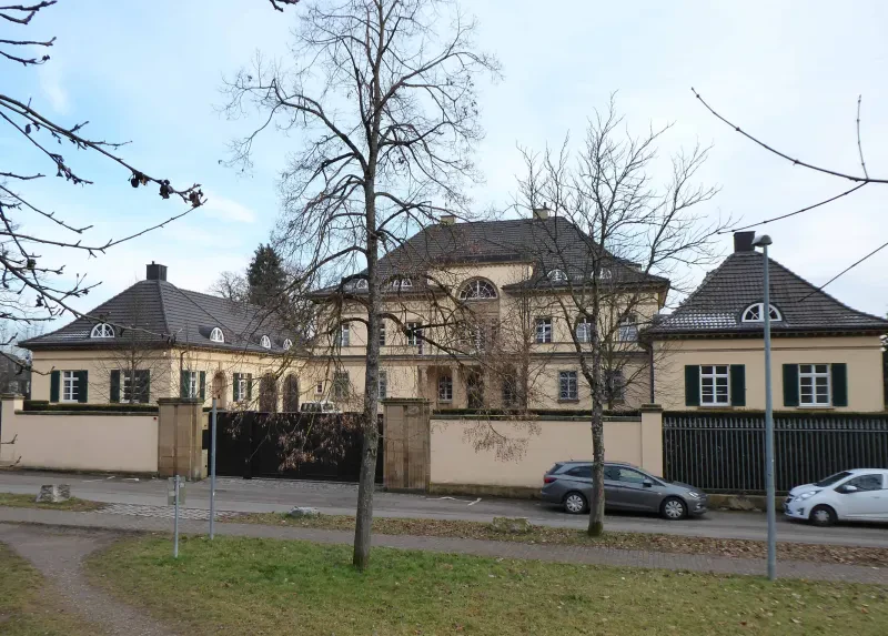 Eine Villa hinter einer Mauer, davor parken Autos.