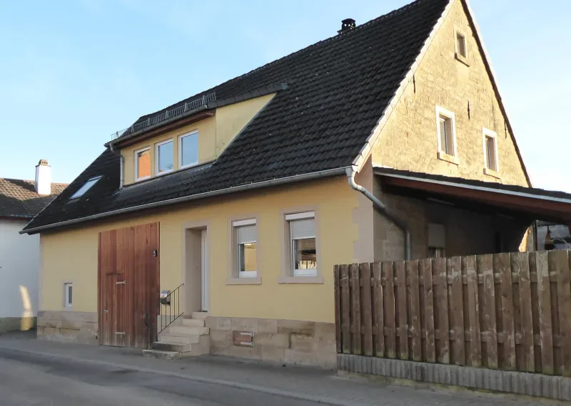 Ein kleines gelb gestrichenes haus, rechts ein Holzbretterzaun.