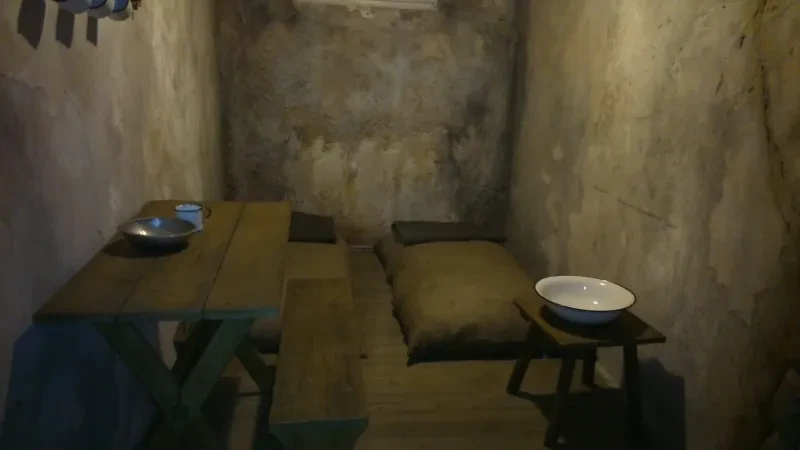 Eine Gefängniszelle mit einem Strohsack, einem Tisch mit Bank, darauf eine Essschüssel und ein Becher sowie einem Hocker mit Waschschüssel.
