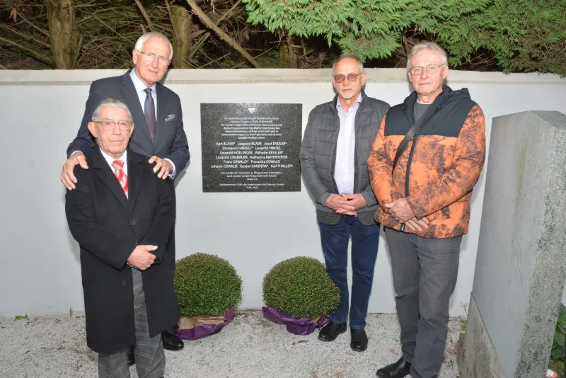 Vier Männer stehen neben einer Gedenktafel auf einem Friedhof. Vor der Mauer stehen zwei Buchsbaumbüsche. Das Foto wurde bei Dunkelheit aufgenommen.