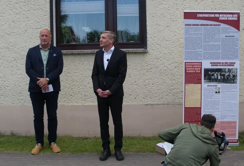 Zwei Männer vor einer Hauswand, rechts von ihnen ein Roll-Up.