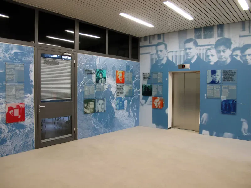 Ausstellungstafel im Foyer eines Hauses, dazwischen eine Tür und eine Fahrstuhltür.
