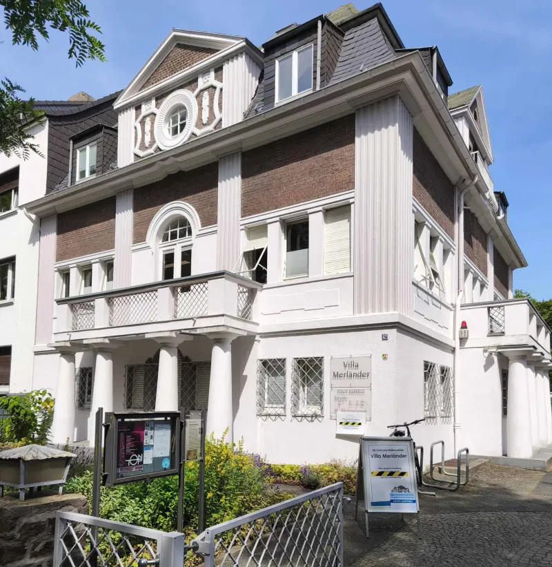 Eine weiße Villa, davor ein Schaukasten und ein Aufsteller. An der Villa ist ein Schild mit der Aufschrift "Villa Merländer" angebracht.